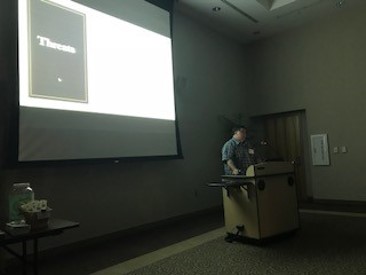 A man in a darkened room, sitting behind a podium, with a projector screen behind him